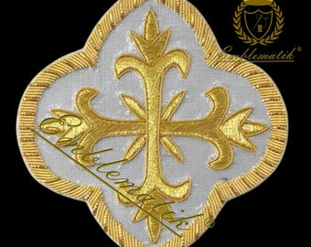May include: A white fabric patch with a gold embroidered cross. The cross has a stylized design with floral details. The patch is surrounded by a gold border with a decorative pattern.
