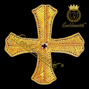 Peut inclure: Un emblème en forme de croix de couleur or avec des broderies complexes. La croix présente une pierre précieuse centrale rouge foncé et est placée sur un fond noir. L'emblème comporte un logo dans le coin supérieur droit.