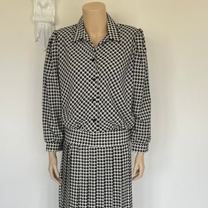 May include: A vintage black and white houndstooth print dress. The dress features a collared button-down top with long sleeves and a pleated skirt. The dress is displayed on a mannequin.