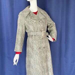 May include: A long-sleeved trench coat featuring a grey and white snakeskin pattern. The coat has a wide collar, a double-breasted button front, and a matching belt with a rectangular buckle. It is worn over a red garment with long sleeves.