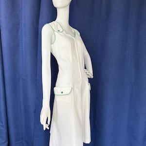 Peut inclure: Robe blanche avec garnitures et accents verts. La robe pr&eacute;sente une encolure en V, une fermeture &eacute;clair sur le devant et deux poches avant. Les garnitures et les accents sont de couleur vert vif. La robe est pr&eacute;sent&eacute;e sur un mannequin.