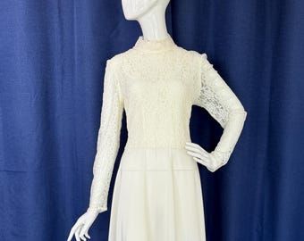 May include: Ivory-colored vintage dress with a high neckline and long sleeves. The bodice features delicate lace detailing, while the skirt flows to a mid-calf length. The dress is displayed on a mannequin against a blue backdrop.