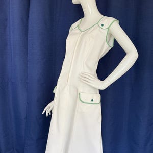 Peut inclure: Robe blanche sans manches avec garniture verte. La robe a une fermeture &eacute;clair sur le devant, deux poches avant et des boutons d&eacute;coratifs verts. La garniture verte souligne l'encolure, les emmanchures et les poches. La robe est de longueur genou et de style r&eacute;tro.