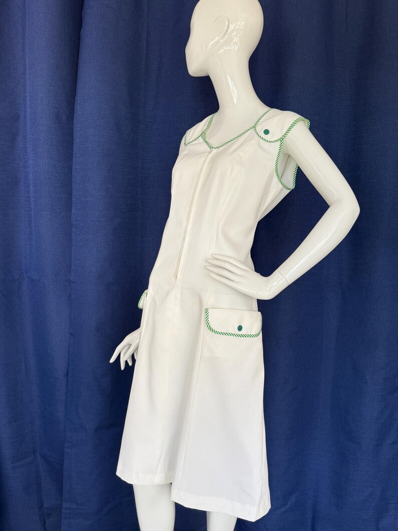 Peut inclure: Robe blanche sans manches avec garniture verte. La robe a une fermeture &eacute;clair sur le devant, deux poches avant et des boutons d&eacute;coratifs verts. La garniture verte souligne l'encolure, les emmanchures et les poches. La robe est de longueur genou et de style r&eacute;tro.