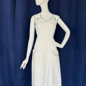 Peut inclure: Robe blanche sans manches avec garniture verte. La robe a une fermeture &eacute;clair sur le devant, deux poches avant et une garniture verte autour de l'encolure et des emmanchures. La robe est pr&eacute;sent&eacute;e sur un mannequin.