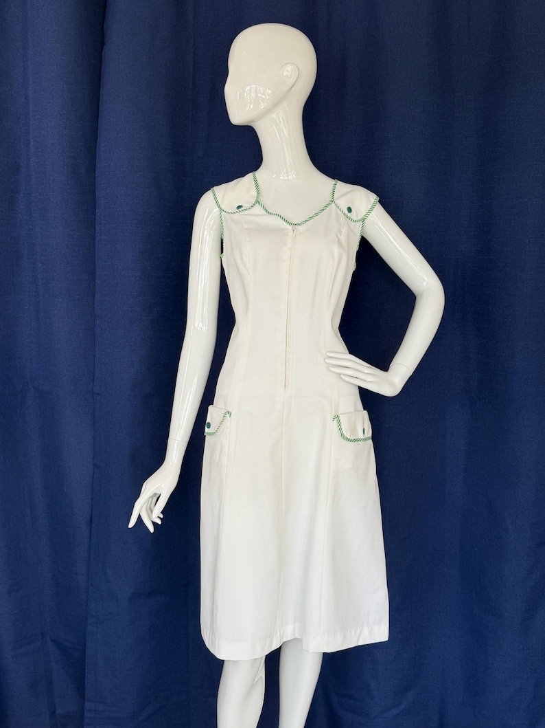 Peut inclure: Robe blanche sans manches avec garniture verte. La robe a une fermeture &eacute;clair sur le devant, deux poches avant et une garniture verte autour de l'encolure et des emmanchures. La robe est pr&eacute;sent&eacute;e sur un mannequin.