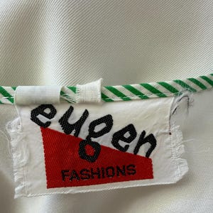 Peut inclure: Gros plan sur une &eacute;tiquette de v&ecirc;tement blanche avec le texte noir "eugen" et le texte rouge "FASHIONS". L'&eacute;tiquette est attach&eacute;e &agrave; un v&ecirc;tement blanc avec une bordure ray&eacute;e verte et blanche.