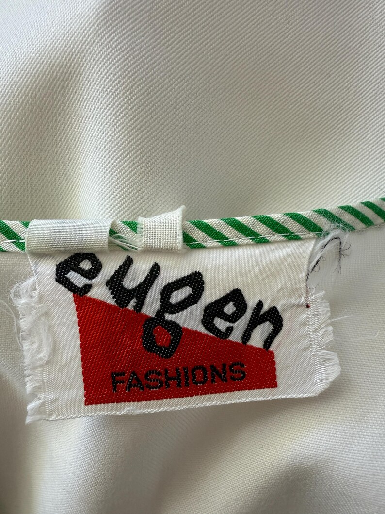 Peut inclure: Gros plan sur une &eacute;tiquette de v&ecirc;tement blanche avec le texte noir "eugen" et le texte rouge "FASHIONS". L'&eacute;tiquette est attach&eacute;e &agrave; un v&ecirc;tement blanc avec une bordure ray&eacute;e verte et blanche.