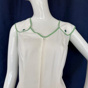 Peut inclure: Robe blanche sans manches avec garniture et boutons verts. La robe a une encolure festonn&eacute;e et une fermeture &eacute;clair sur toute la longueur. La garniture verte est un motif de corde torsad&eacute;e. La robe est pr&eacute;sent&eacute;e sur un mannequin.