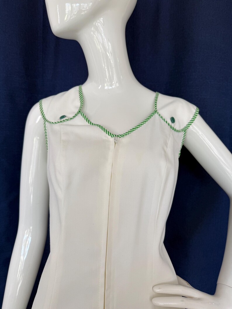 Peut inclure: Robe blanche sans manches avec garniture et boutons verts. La robe a une encolure festonn&eacute;e et une fermeture &eacute;clair sur toute la longueur. La garniture verte est un motif de corde torsad&eacute;e. La robe est pr&eacute;sent&eacute;e sur un mannequin.