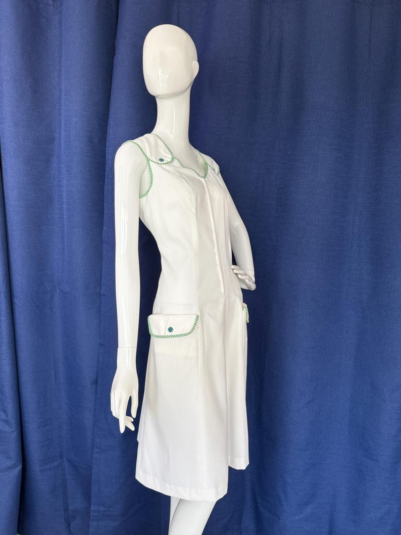 Peut inclure: Robe blanche avec garnitures et accents verts. La robe pr&eacute;sente une encolure en V, une fermeture &eacute;clair sur le devant et deux poches avant. Les garnitures et les accents sont de couleur vert vif. La robe est pr&eacute;sent&eacute;e sur un mannequin.