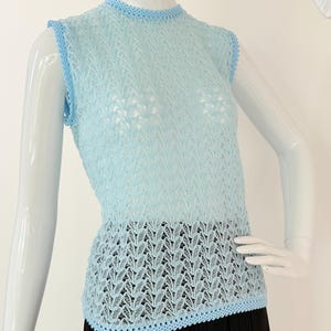 May include: Light blue sleeveless top with a delicate openwork design. The top features a round neckline and armholes trimmed with a lighter shade of blue. The bottom hem is also trimmed with the same lighter blue color. The top is displayed on a mannequin.