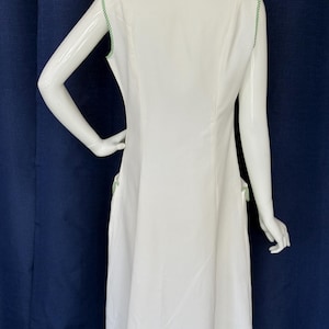 Peut inclure: Robe blanche sans manches avec une bordure verte le long de l'encolure et des emmanchures. La robe a une silhouette simple en forme de A et des bordures vertes sur les poches. La robe est pr&eacute;sent&eacute;e sur un mannequin.