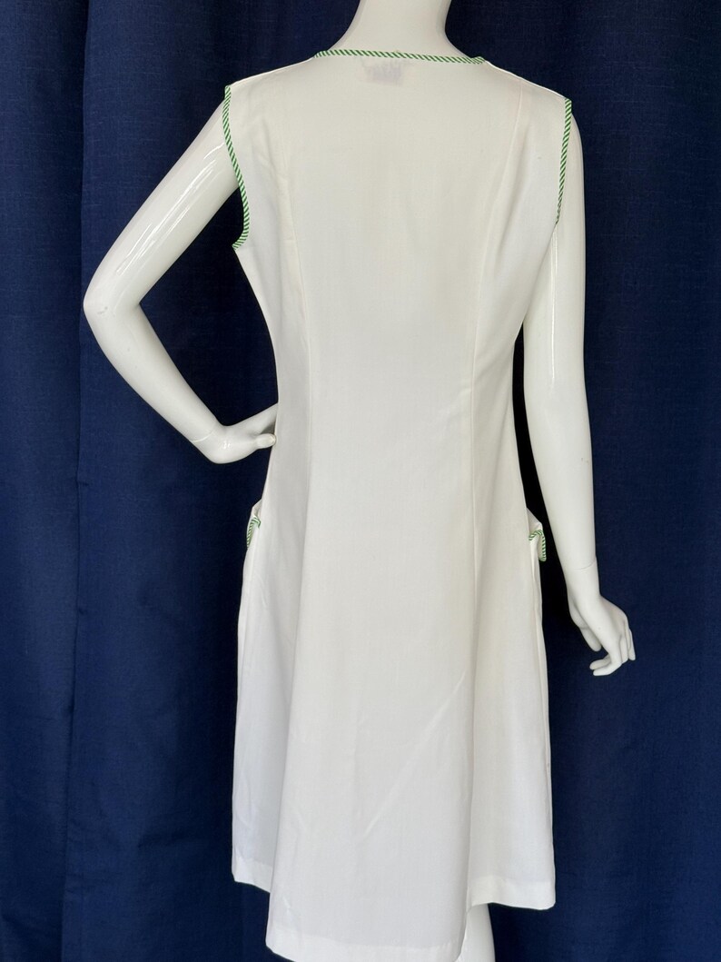 Peut inclure: Robe blanche sans manches avec une bordure verte le long de l'encolure et des emmanchures. La robe a une silhouette simple en forme de A et des bordures vertes sur les poches. La robe est pr&eacute;sent&eacute;e sur un mannequin.