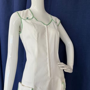 Peut inclure: Robe blanche sans manches avec garniture et d&eacute;tails de poches verts. La robe pr&eacute;sente une fermeture &eacute;clair sur le devant et des boutons verts. La garniture est un motif de corde verte et blanche. La robe est pr&eacute;sent&eacute;e sur un mannequin blanc.