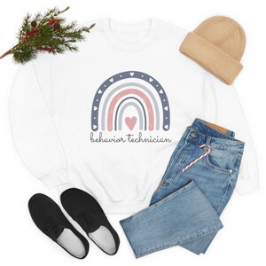 May include: White sweatshirt with a rainbow design in shades of pink, blue, and gray. The rainbow has a heart in the center and the text "behavior technician" below it.
