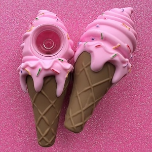 May include: Two novelty ice cream cone-shaped objects. The cones are brown with a waffle pattern. The ice cream is pink with colorful sprinkles. One has a circular opening at the top. The background is a pink, glittery surface.