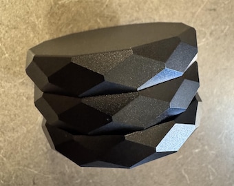 Puede incluir: Una pila de tres posavasos circulares de color negro mate, cada uno con un diseño de borde geométrico facetado. Los posavasos son uniformes en color y forma, con un acabado de superficie texturizado que capta la luz a lo largo del perímetro angular y multifacético.