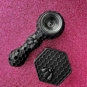 May include: Black glass pipe with a honeycomb design and a bee accent. The pipe is paired with a matching hexagonal tray, also featuring a honeycomb pattern and a bee. The items are set against a pink glitter background.