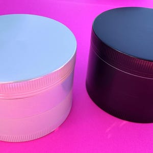 May include: Two cylindrical herb grinders, one in a metallic silver finish and the other in a matte black finish. Both grinders feature a multi-part design with a textured, ribbed grip along the edges of the lid and the base sections.