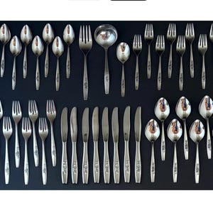 May include: A complete set of vintage silverware, including spoons, forks, knives, and a serving spoon, arranged on a dark surface. The silverware has a floral design on the handles and a polished finish.