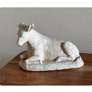 May include: A white, weathered-looking sculpture of a reclining animal, possibly a calf or lamb. The sculpture is resting on a rectangular base and has a textured surface. The piece is likely a decorative item.