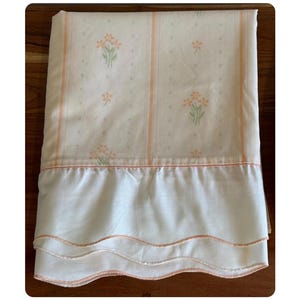 May include: Cream-colored bed sheet with a floral pattern and vertical peach stripes. The sheet features a ruffled edge with a scalloped design. The floral pattern includes small orange flowers with green leaves. The sheet is folded on a wooden surface.