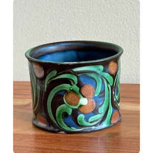 May include: A ceramic cylindrical vase with a dark brown exterior, adorned with green, orange, and blue floral patterns. The interior is a vibrant blue. The vase sits on a wooden surface.