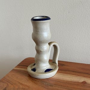 May include: A handmade ceramic candlestick holder. The off-white holder has a blue rim and accents. It features a handle and a circular base with blue details. The candlestick holder is sitting on a wooden surface.