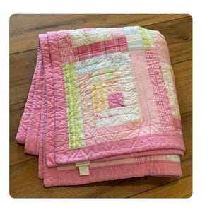 May include: A folded pink and white patchwork quilt with a log cabin design. The quilt features squares of pink, white, and pastel colors, including green and yellow. The edges are a solid pink, and the quilt is folded neatly.