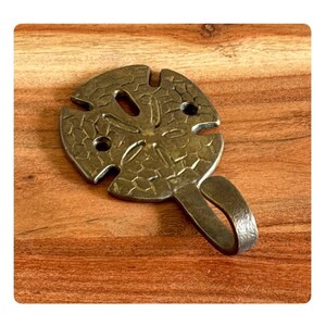 May include: A bronze-colored metal wall hook in the shape of a sand dollar. The hook has a textured surface and a curved hook for hanging items. The sand dollar design features a central star pattern and several small holes.