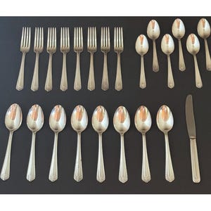 May include: A set of silver-colored flatware, including forks, spoons, and a knife, arranged on a dark surface. The silverware has a decorative pattern on the handles. The set includes multiple forks, spoons, and a single knife, suitable for a formal dining setting.
