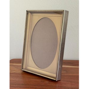 May include: A gold-toned picture frame with an oval opening. The frame has a rectangular shape with a decorative textured edge. The frame is angled on a wooden surface, with a neutral background.
