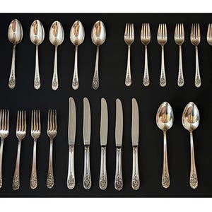May include: A collection of silver-colored flatware, including spoons, forks, and knives, arranged on a black surface. The silverware has decorative floral patterns on the handles. The spoons and knives are placed in parallel rows, while the forks are arranged in perpendicular rows.