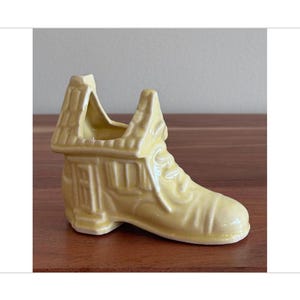 May include: A yellow ceramic shoe planter with a house-shaped top. The shoe has a detailed design and a small door on the side.