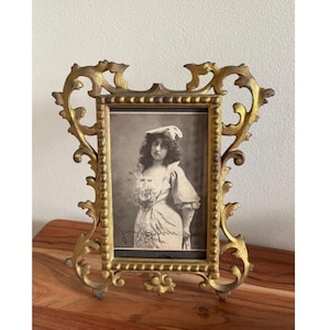 May include: Ornate gold-colored picture frame with a vintage black and white photograph of a woman. The frame has an elaborate, scrolling design and sits on a wooden surface. The photograph features a woman in a dress and hat.