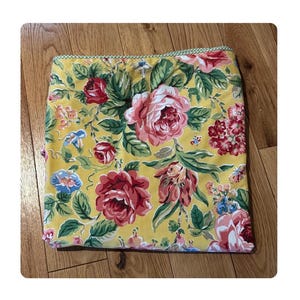 May include: Yellow floral pillow cover with a pattern of large pink and red roses, green leaves, and small blue flowers. The pillow cover has a green and white checkered border.