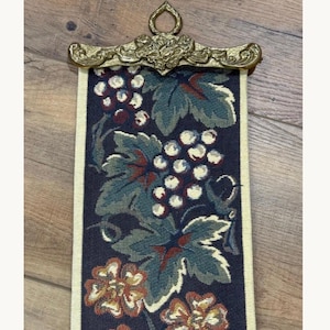 May include: A vertical wall hanging tapestry featuring a dark navy background with a woven pattern of green grape leaves, white grape clusters, and orange flowers. The textile is suspended from an ornate, metallic gold-toned metal hanger.