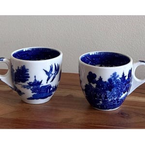 May include: Two white ceramic teacups with blue Willow pattern designs. Each cup has a handle and a blue interior. The cups are placed on a wooden surface.