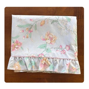 May include: A light blue floral patterned bed sheet with pink, peach, and green flowers. The sheet has a ruffled edge with a thin pink trim. The floral pattern is spread across the sheet.