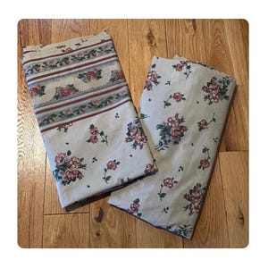May include: Two folded floral patterned bed sheets. The sheets are a light cream color with a repeating pattern of pink and red roses and green leaves. One sheet has a decorative border with a floral and stripe design.