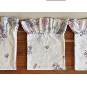 May include: Three off-white fabric valances with a floral pattern in pink, purple, and green. Each valance has a ruffled top and a rectangular body. The valances are arranged on a wooden surface.