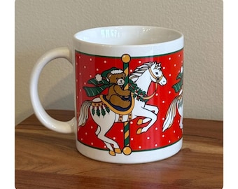 May include: A white ceramic mug with a curved handle. The mug features a red background with white polka dots and a carousel horse design. A brown teddy bear is riding the white horse. The mug is sitting on a wooden surface.