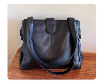 May include: Black leather shoulder bag with a flap closure and a long adjustable strap. The bag has a rectangular shape with a rounded flap and a stitched design. The bag is sitting on a wooden surface.