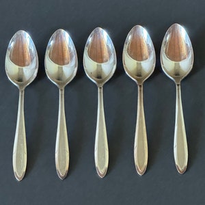 May include: Five silver spoons with ornate handles. The spoons are arranged in a row on a dark gray surface.