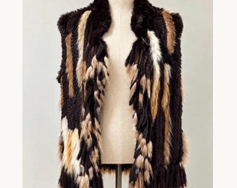 May include: A sleeveless vest made of multi-colored fur panels in shades of dark brown, tan, and white. The garment features a textured, layered appearance with vertical strips of fur arranged in a pattern, a wide collar, and an open front design.