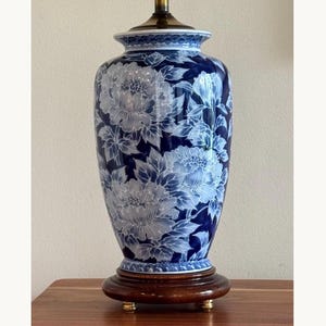 May include: A blue and white ceramic table lamp base featuring a detailed floral pattern of large peony blossoms and leaves. The cylindrical vessel sits on a circular dark wood pedestal base with small metallic feet and a brass hardware top.