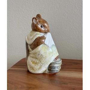 May include: A ceramic figurine of a brown squirrel wearing a cream-colored shawl and a light yellow dress. The squirrel is holding a white object and sitting on a small, gray basket. The figurine is on a wooden surface.