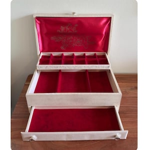 May include: A vintage jewelry box with a cream-colored exterior and a vibrant red velvet interior. The box features multiple compartments and a drawer for storing jewelry. The interior lid has a gold floral design.