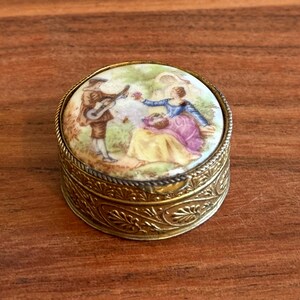 May include: A round metal trinket box with a gold-toned finish and intricate embossed leaf patterns on the sides. The lid features a circular porcelain inlay depicting a colorful scene of a person playing a guitar for another person sitting nearby.